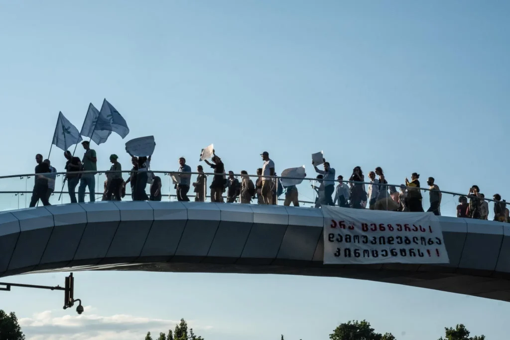 A group of people holding signs and flags marches across a modern pedestrian bridge, highlighting the movement for taming culture in Georgia. A large banner with Georgian text hangs on the side under a clear blue sky.
