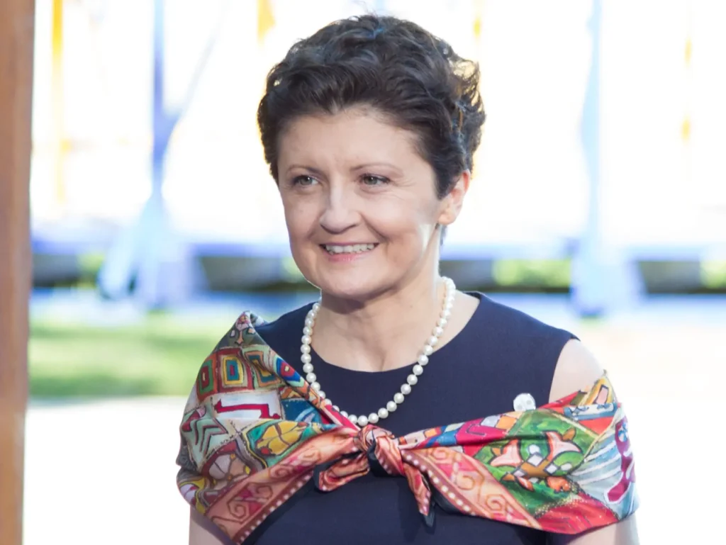 A middle-aged woman with short brown hair smiles, wearing a colorful patterned scarf draped over her shoulders, a dark dress, and a pearl necklace, embodying the warmth and tradition of taming culture in Georgia. The softly blurred background hints at outdoor sunlight.