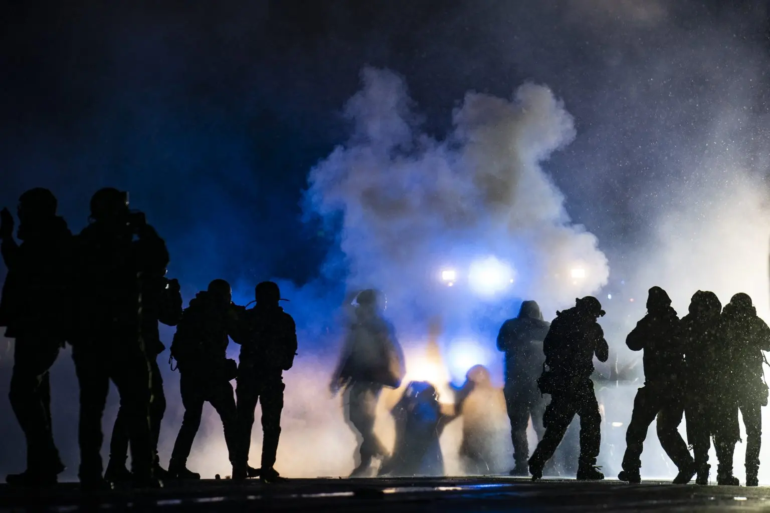 Silhouetted people stand and move in front of heavy smoke and bright lights at night, suggesting a protest or chaotic scene possibly sparked by recent anti-protest bills. Blue and white lighting creates a dramatic atmosphere.