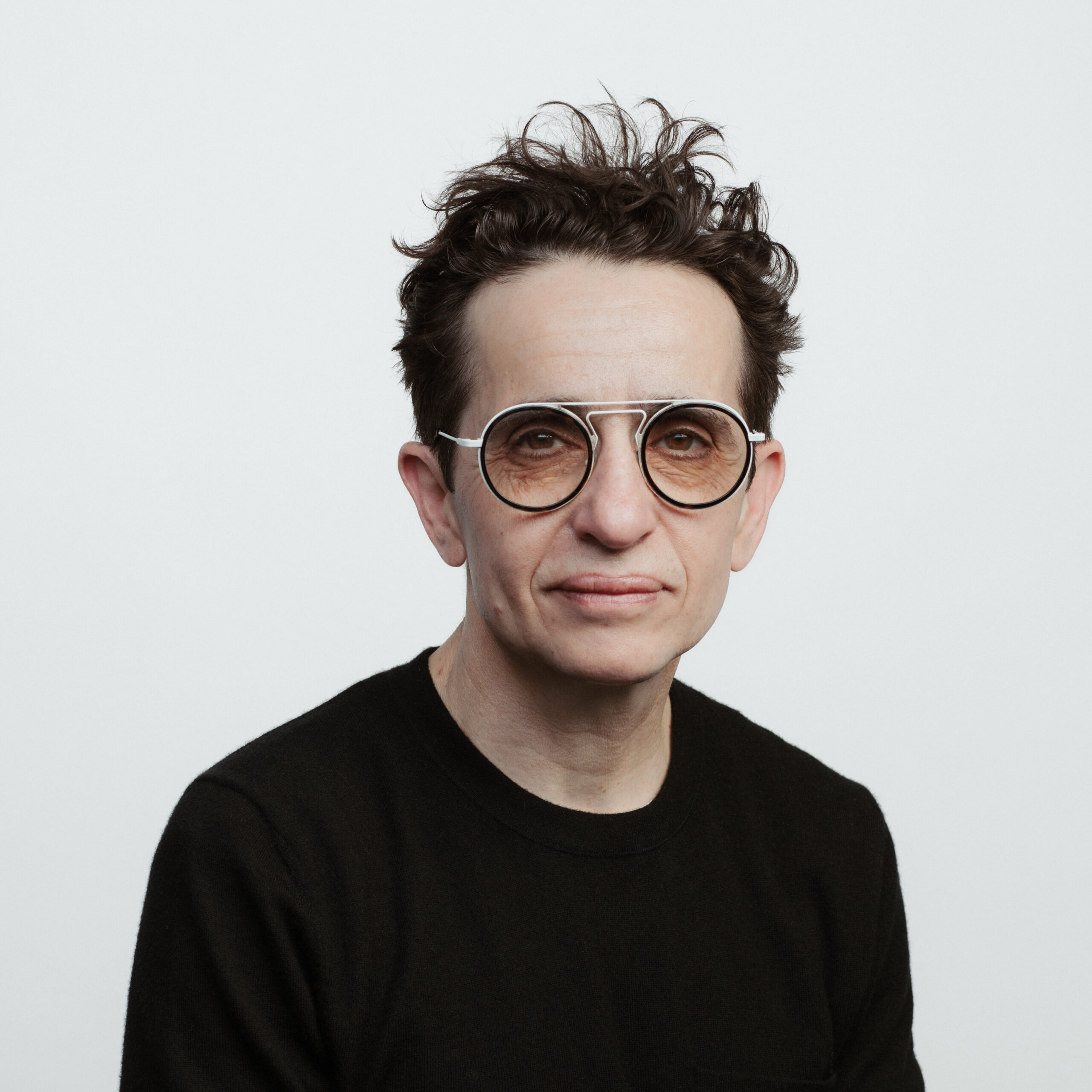 A person with short, tousled dark hair wearing large round glasses and a black sweater, poses against a plain white background, looking directly at the camera with a neutral expression.