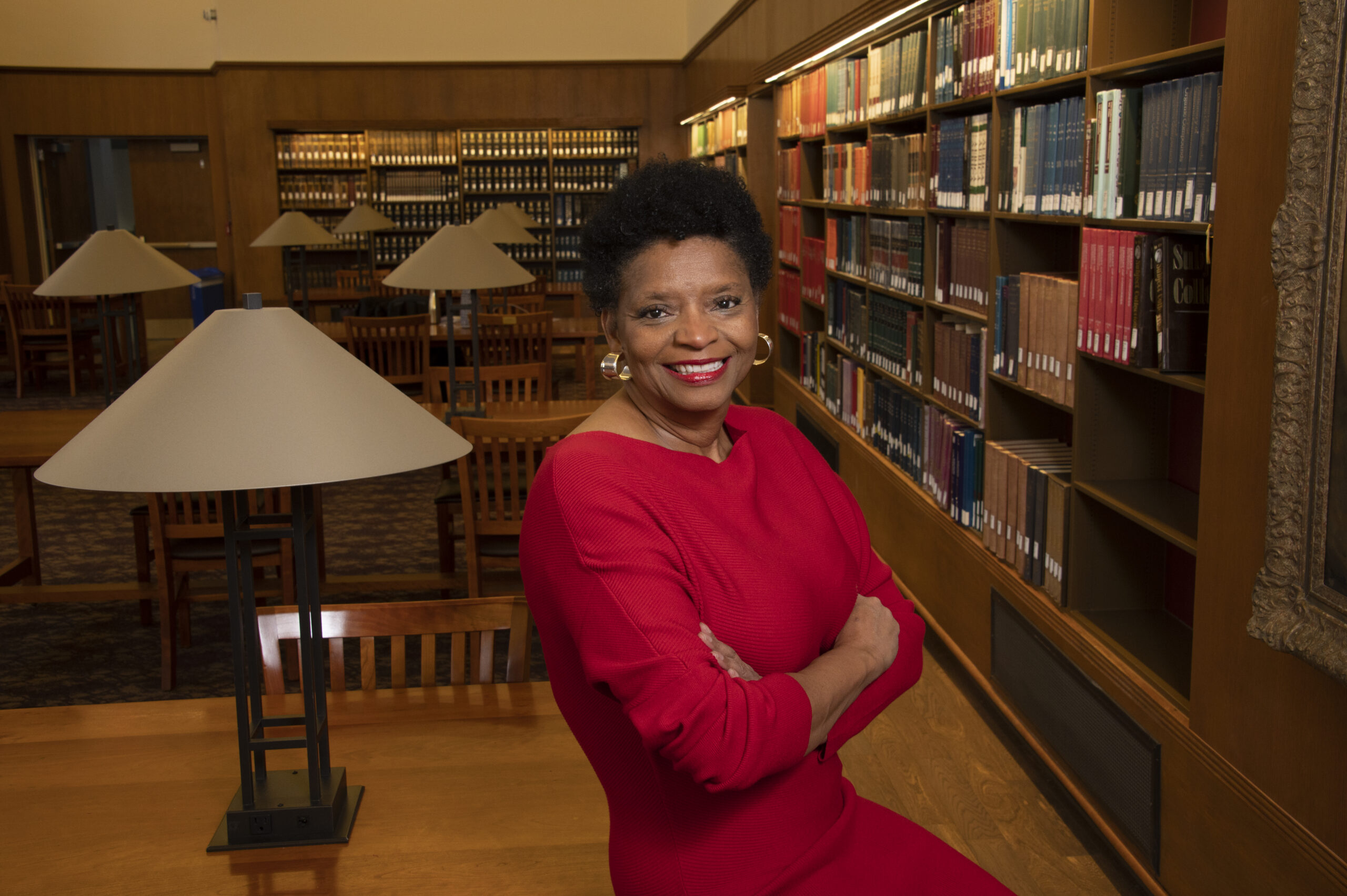 A woman in a red dress smiles with arms crossed, standing by a wooden table in a library filled with bookshelves, lamps, and wooden chairs.
