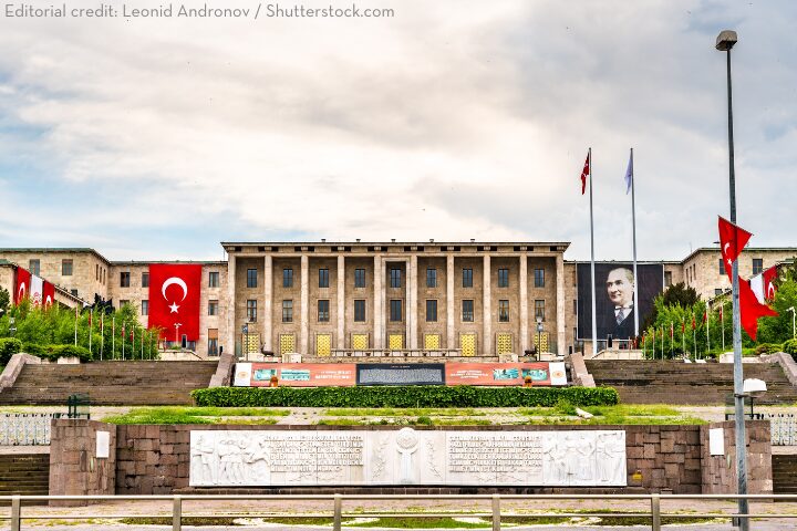 The Grand National Assembly of Turkey building in Ankara, decorated with Turkish flags and a large portrait of Mustafa Kemal Atatürk, with manicured greenery and stone carvings in the foreground.