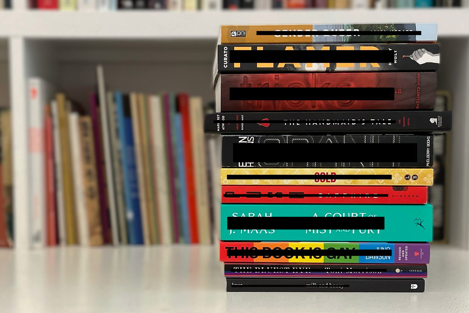 A stack of colorful books lies on a white surface in front of a blurred bookshelf, their spines displaying various titles and colors. This vibrant scene highlights the importance of accessible Book Ban Resources for every reader.