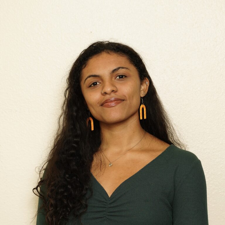 A woman with long curly hair wearing a dark green top and orange earrings stands in front of a plain light-colored wall, smiling softly at the camera.
