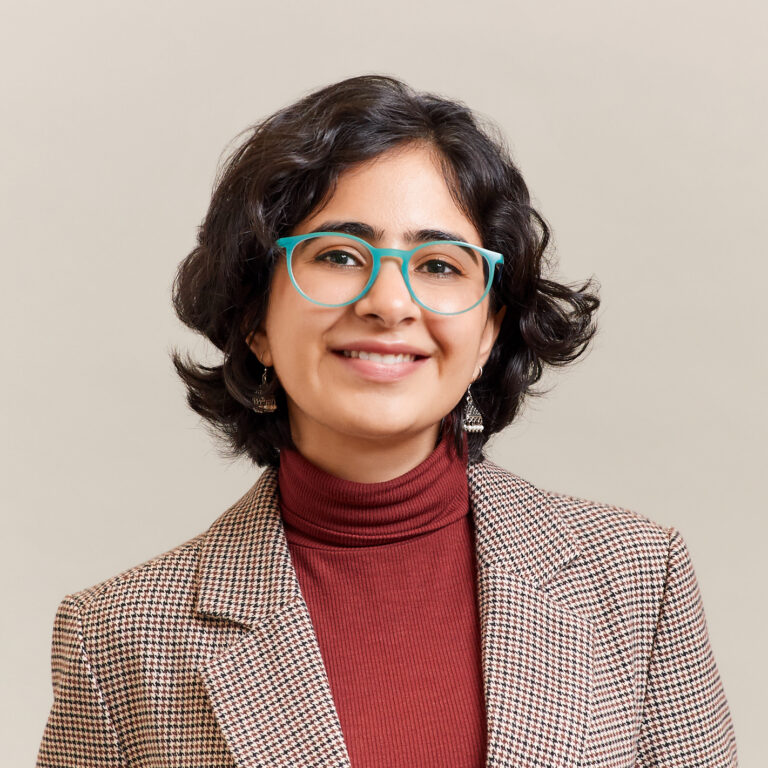 A person with short, dark, wavy hair wears teal glasses, dangling earrings, a brown turtleneck, and a brown checked blazer, smiling in front of a plain beige background.