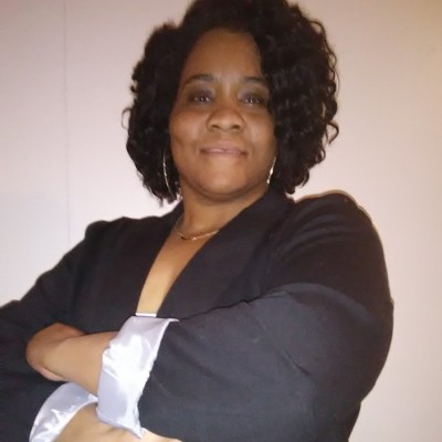 A woman with curly hair smiles confidently at the camera, wearing a dark blazer with light-colored cuffs and her arms crossed. The background is plain and light-colored.