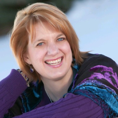 A woman with short, light brown hair smiles at the camera. She is wearing a purple sweater and earrings, with a blurred outdoor background.
