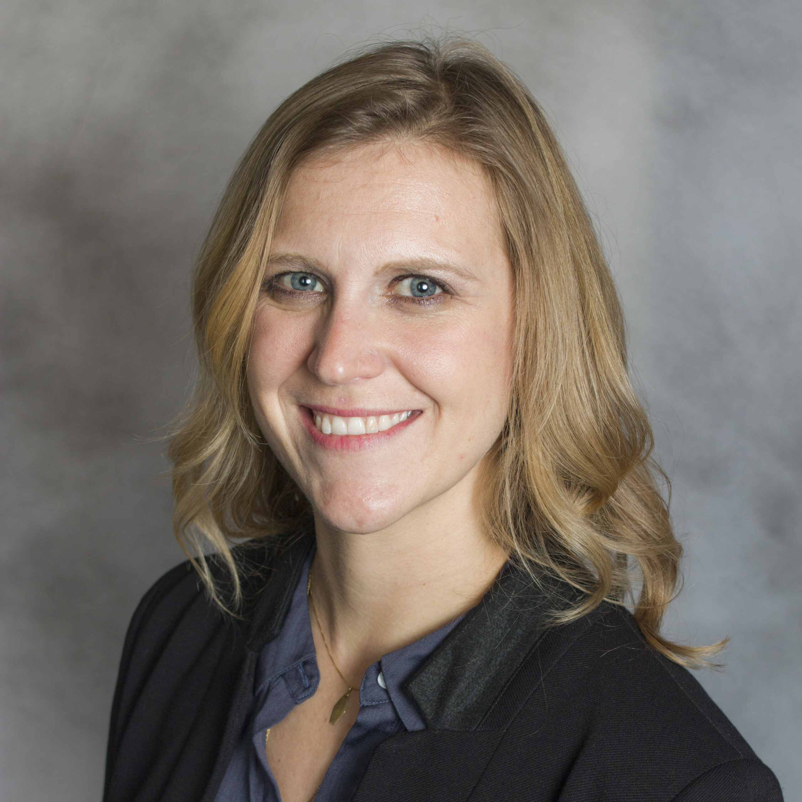 A woman with wavy blonde hair, wearing a dark blazer and blue blouse, smiles at the camera against a neutral, softly blurred background.