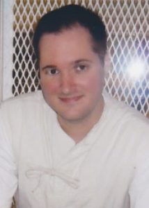 A man with short dark hair, wearing a white shirt, sits and smiles slightly. Behind him is a white metal mesh or grille background, suggesting a thoughtful moment—perhaps related to his Writing for Justice Fellowship.