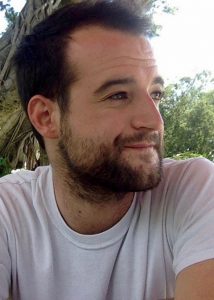 A man with short dark hair and a beard wearing a white t-shirt sits outdoors, smiling slightly and looking to his left, thoughtfully reflecting on his Writing for Justice Fellowship amidst greenery and tree branches in the background.