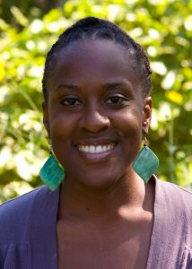 A woman with braided hair smiles at the camera, wearing large green square earrings and a purple top, with a leafy green background—radiating confidence akin to a Writing for Justice Fellowship recipient.