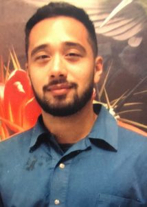 A man with short black hair and a beard, wearing a blue collared shirt, stands in front of a colorful abstract background featuring red and yellow shapes, representing the spirit of the Writing for Justice Fellowship.