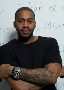 A man with a beard and tattooed arms stands with his arms crossed, wearing a black t-shirt and a watch, in front of a whiteboard filled with notes from the Writing for Justice Fellowship.
