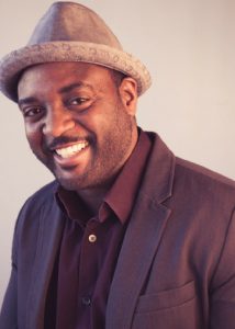 A man wearing a light gray fedora, dark blazer, and maroon shirt smiles at the camera against a plain light background, embodying the confidence of a Writing for Justice Fellowship recipient.