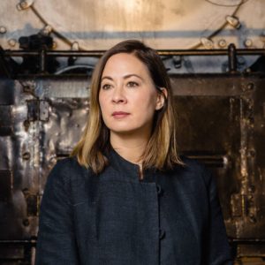 A woman with shoulder-length light brown hair, wearing a dark jacket, stands in front of a large industrial metal background, looking slightly to the side with a neutral expression.