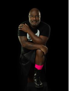A man poses against a black background, smiling and resting one arm on his raised knee. He wears a black t-shirt and shorts, with a bright pink sock visible on his lower leg.