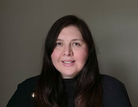 A woman with long dark hair smiles softly at the camera against a plain, neutral background. She is wearing a dark top.