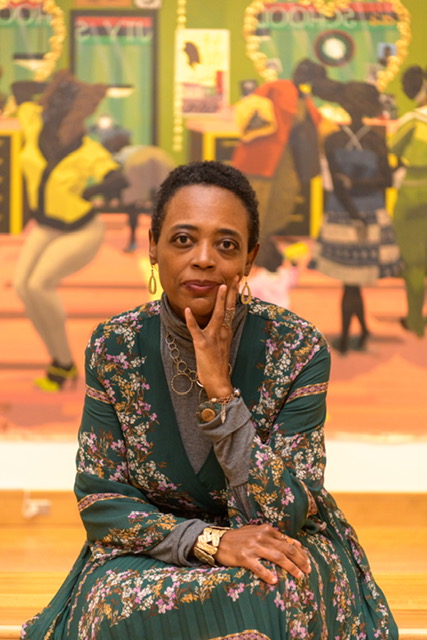 A woman with short hair and hand resting on her face sits in front of a colorful, blurred background. She wears a green floral jacket, gold earrings, and a necklace, looking directly at the camera.