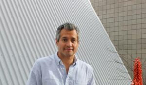 A man wearing a light blue button-up shirt stands in front of a corrugated metal wall and grid-patterned building exterior.