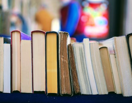 A row of books stands upright on a table, their colorful spines and worn pages hinting at poets across lines. The background is blurred with indistinct shapes and colors.