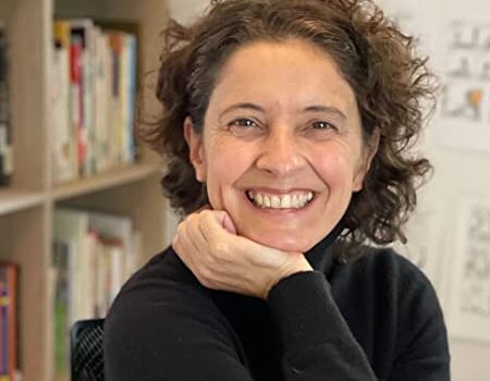 A woman with curly brown hair, wearing a black top, smiles warmly with her chin resting on her hand. She is sitting indoors, with bookshelves and papers visible in the background.