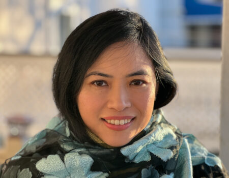 A woman with black hair smiles at the camera, wearing a dark shawl with light blue floral patterns. The background is softly blurred with warm lighting.