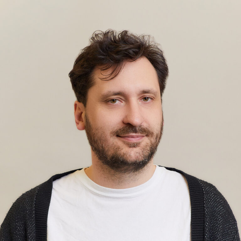 A man with wavy brown hair and a beard, wearing a white T-shirt and a dark cardigan, stands against a plain light-colored background, looking at the camera with a slight smile.