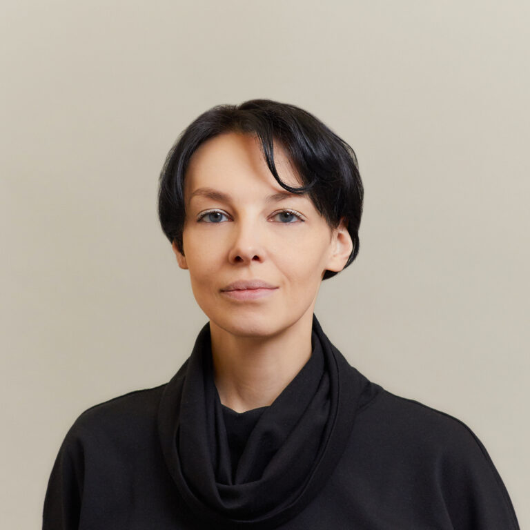 A woman with short dark hair wearing a black top with a loose collar stands against a plain light gray background, looking calmly at the camera.