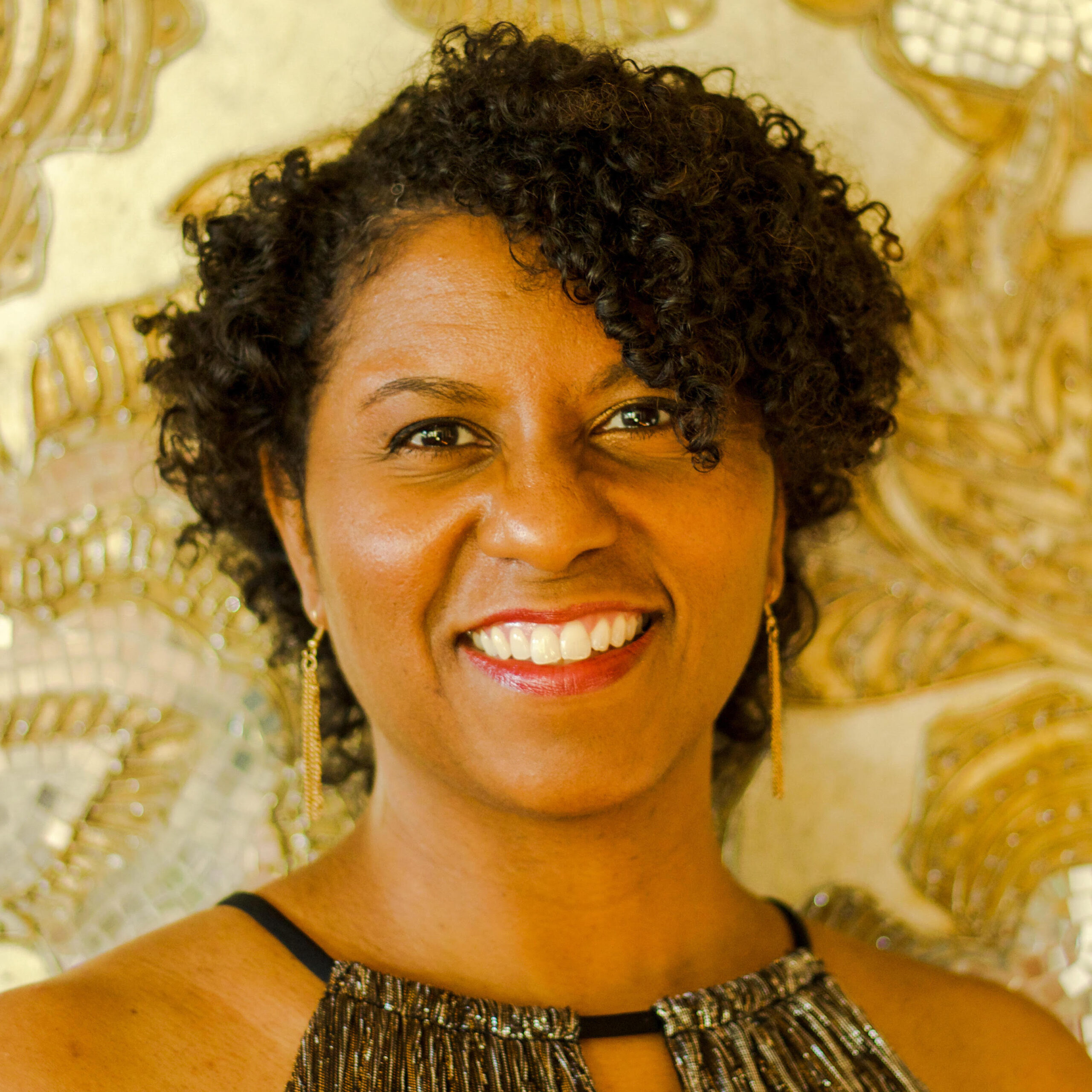 Smiling woman with curly hair, wearing a sleeveless top and dangling earrings, stands in front of a decorative gold floral background.