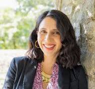 A woman with shoulder-length dark hair and gold hoop earrings smiles while leaning against a stone wall. She is wearing a black blazer over a pink patterned blouse. The background is out of focus, showing greenery and light.