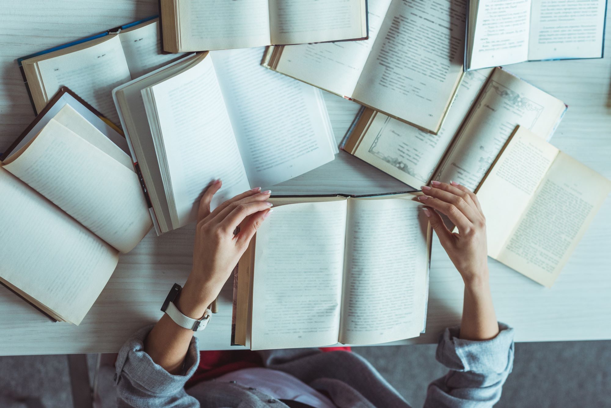 A person sits at a table covered with open books, holding one book and reading. The scene suggests studying or research, with pages spread out around their hands on a light-colored surface.