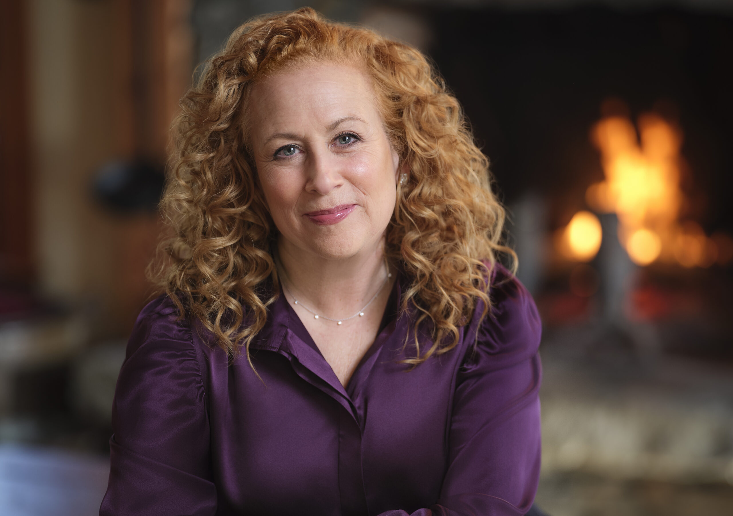 A woman with curly red hair wearing a purple blouse smiles softly at the camera, seated indoors with a lit fireplace in the blurred background.