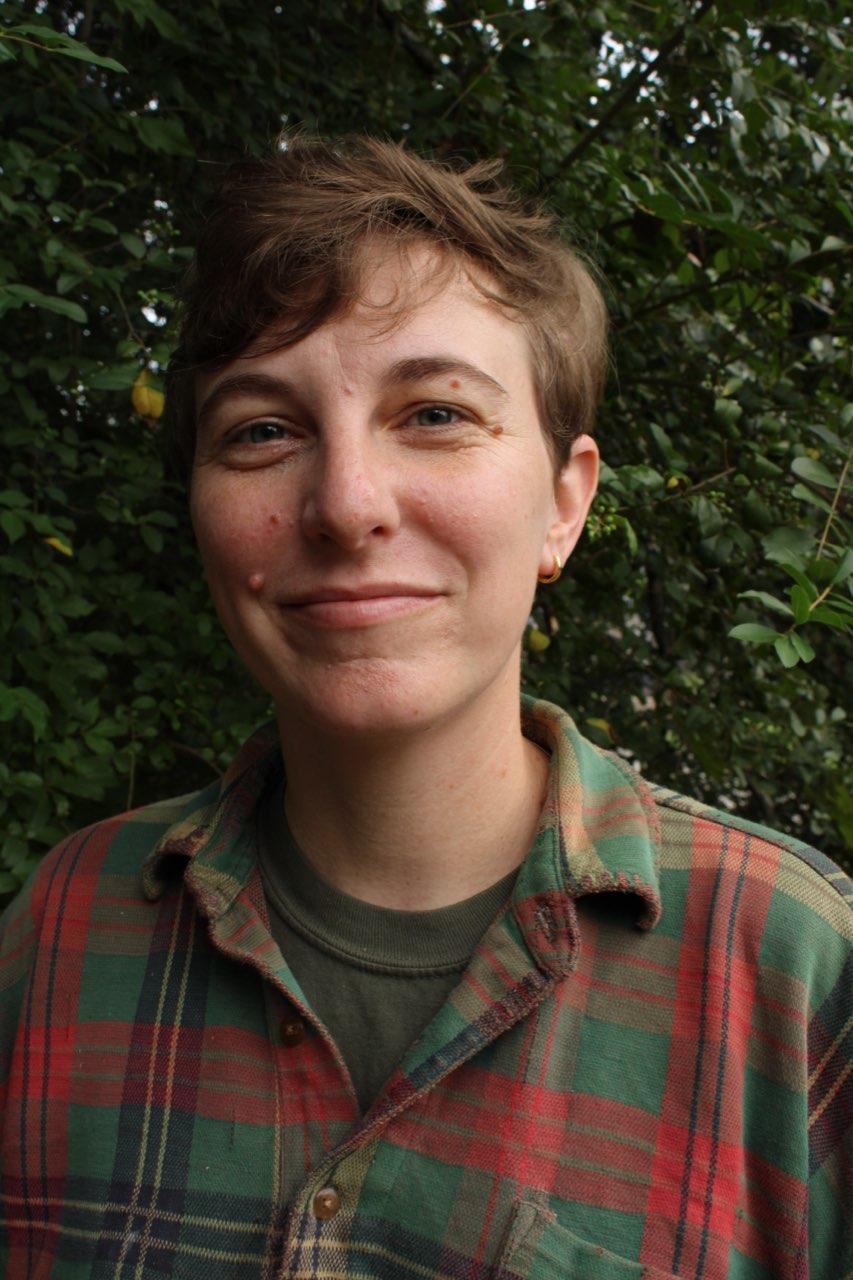 A person with short brown hair, wearing a green and red plaid shirt over a dark t-shirt, smiles gently while standing in front of leafy green trees.