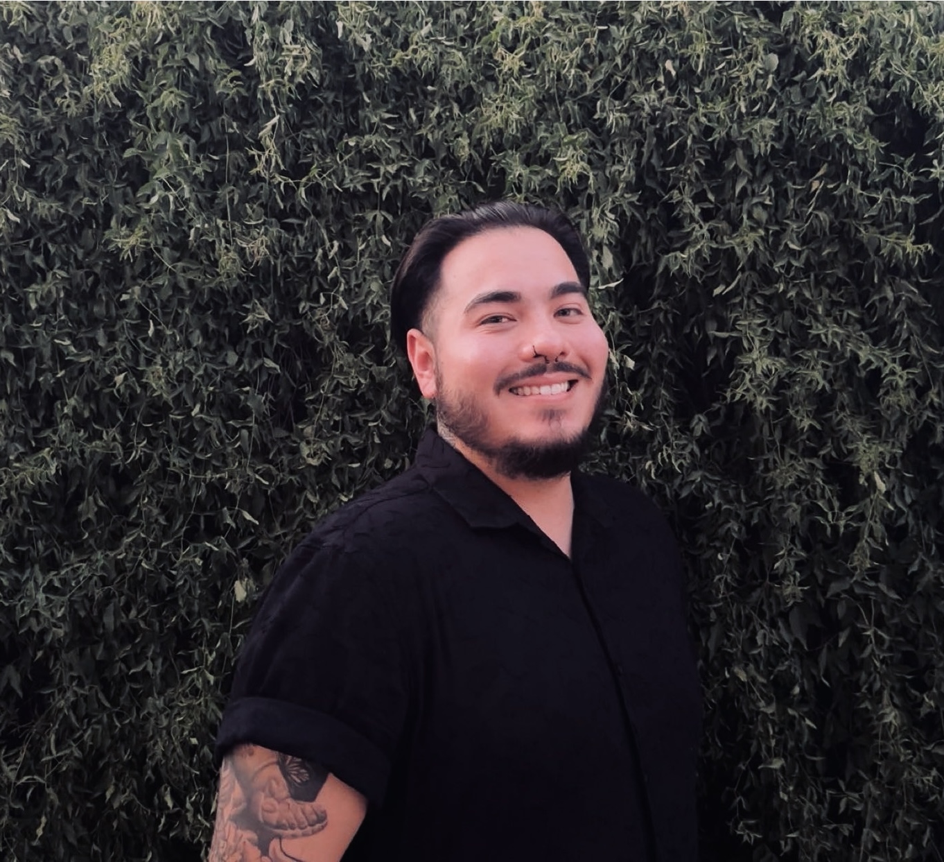 A smiling person with dark hair, a beard, and a tattoo on their arm stands outdoors in front of a dense green leafy background, wearing a short-sleeved black shirt.