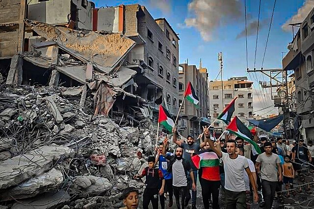 A group of people wave Palestinian flags and march beside a large pile of rubble from destroyed buildings in an urban area, with damaged structures visible in the background under a partly cloudy sky.