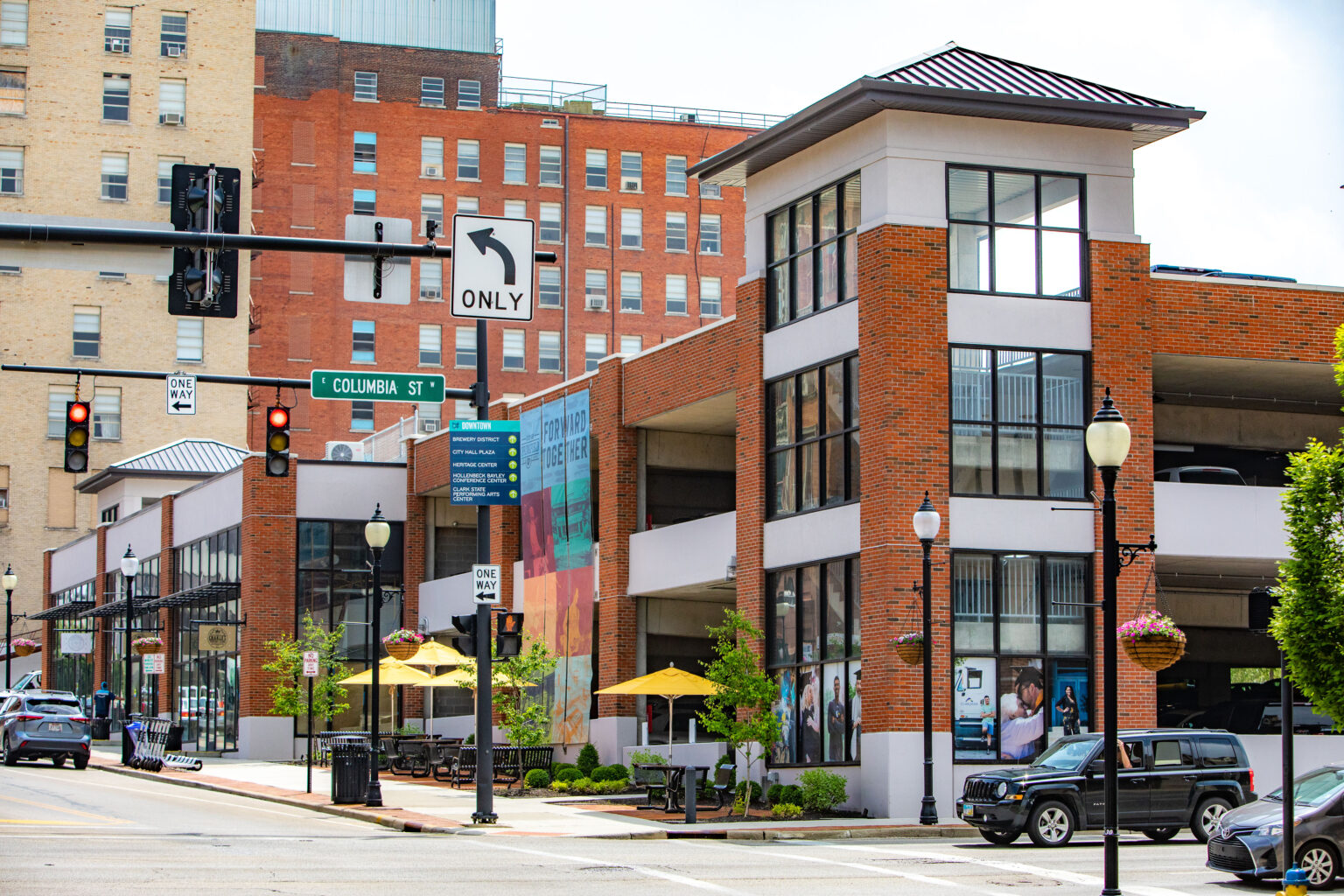 A busy urban street corner with a brick parking garage, colorful mural, street signs, cars parked along the road, and outdoor seating under yellow umbrellas on the sidewalk.