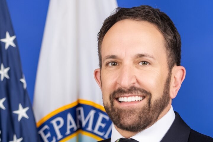 A man with short dark hair and a beard, wearing a suit and tie, smiles in front of a U.S. flag and a white flag with a partial government seal visible on a blue background.