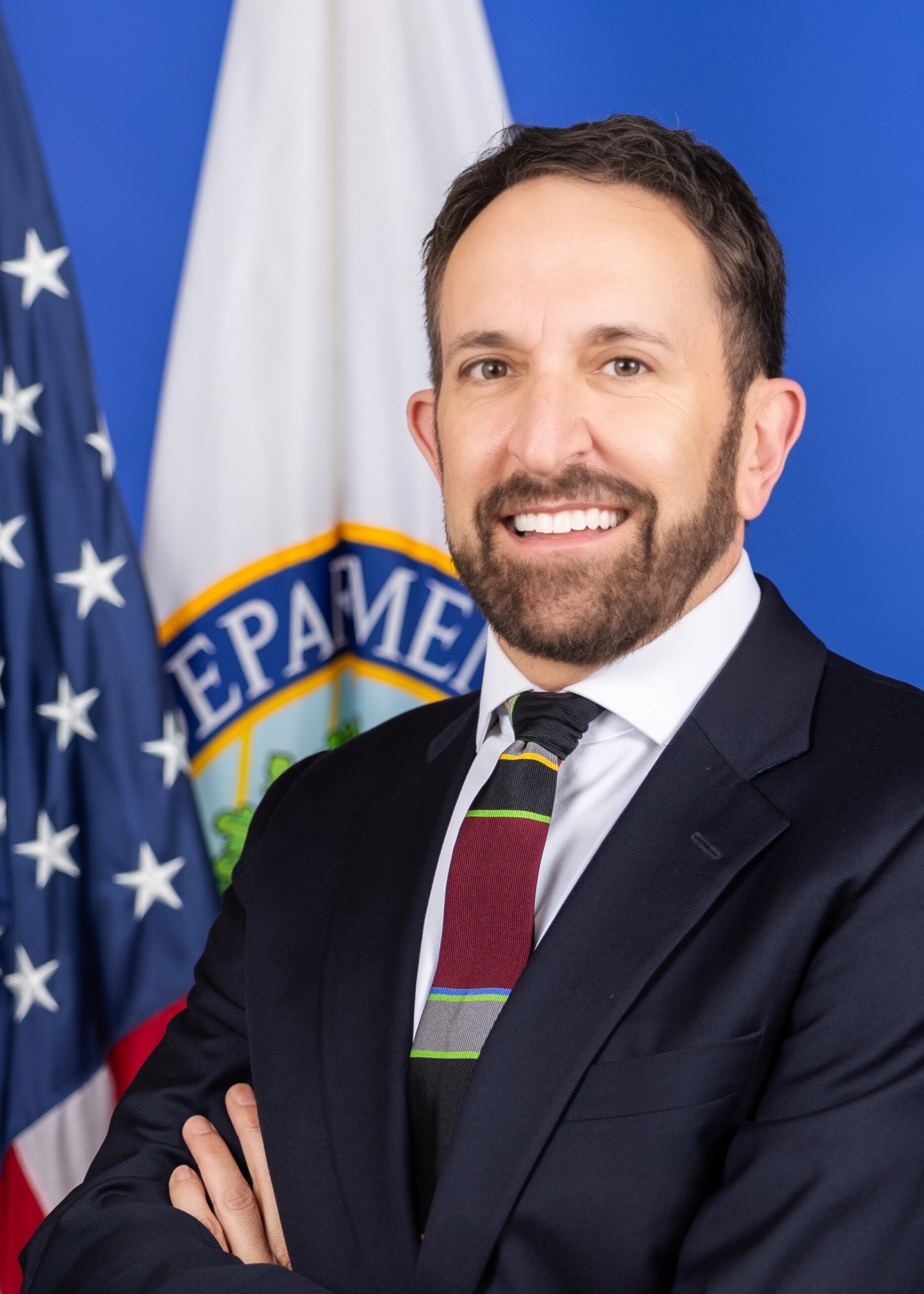 A man in a suit and striped tie stands smiling with arms crossed in front of U.S. and Department of Education flags, against a blue background.