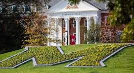 A large M made of flowers on a grassy slope in front of a brick building with white columns. The building has a red door, and trees surround the area.