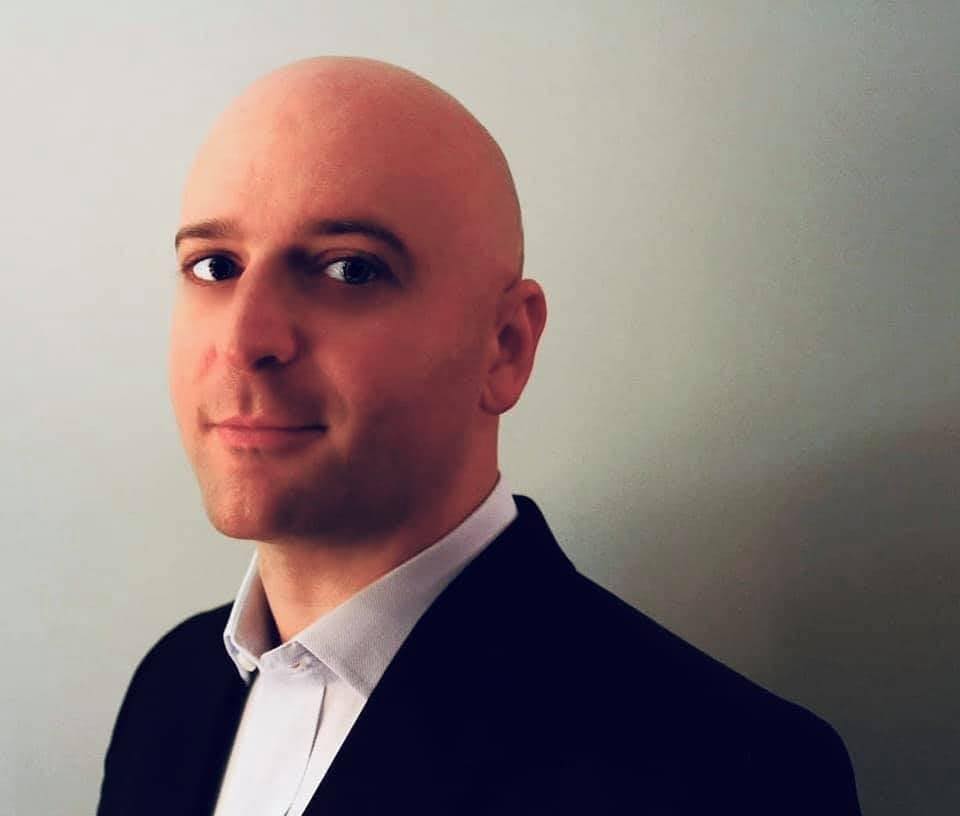 A bald man wearing a dark suit jacket and a white collared shirt stands against a plain light-colored background, looking at the camera with a slight smile.