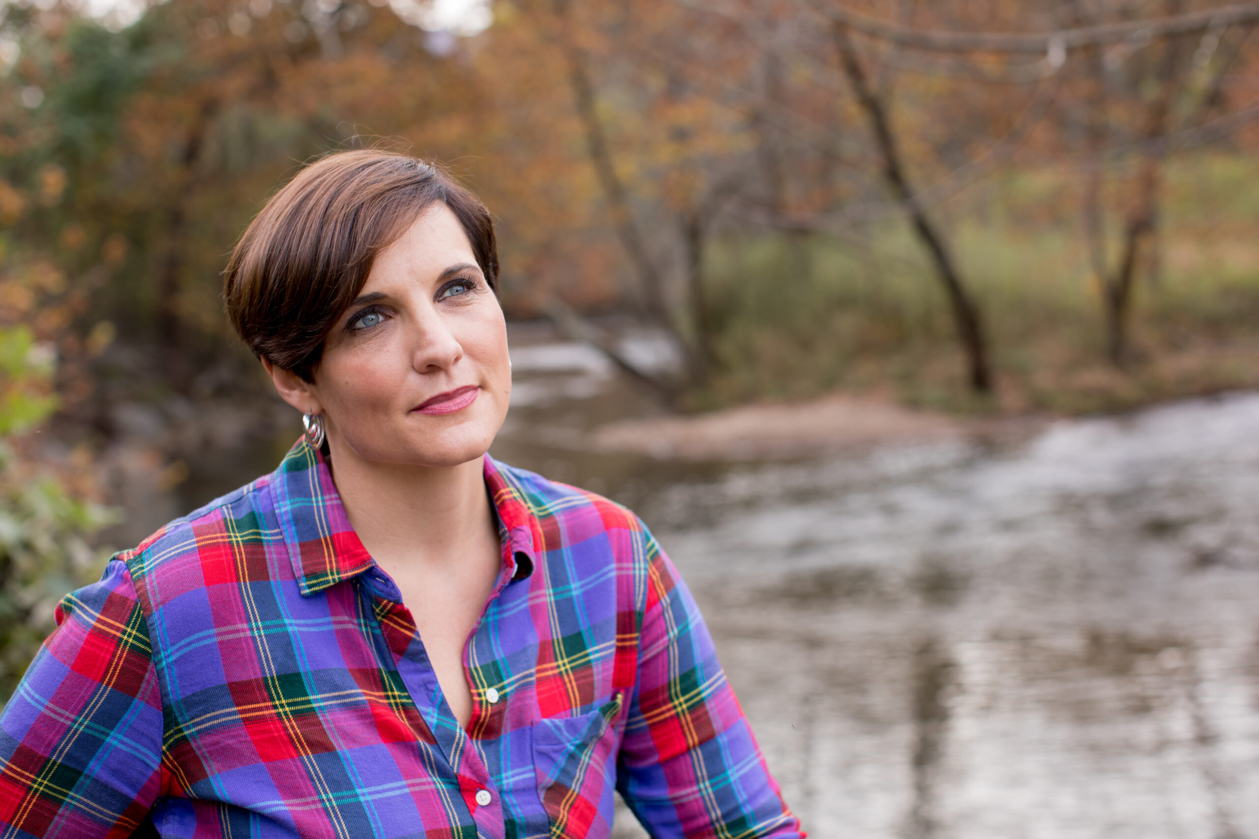 A woman with short brown hair in a colorful plaid shirt stands outdoors by a creek, gazing thoughtfully upward. The background features trees with autumn leaves and gently flowing water.