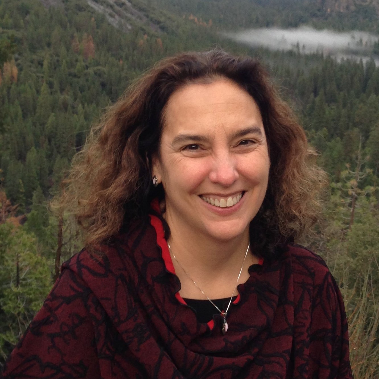 A woman with wavy brown hair smiles outdoors, wearing a dark red and black patterned top and a necklace. Behind her is a forested landscape with tall trees and mist in the distance.