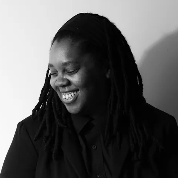 A person with long dreadlocks, wearing a dark jacket and shirt, smiles while looking down. The photo is black and white with a plain background.