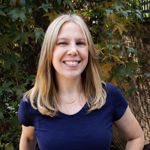 A woman with straight blonde hair, wearing a navy blue shirt, stands outdoors in front of leafy green plants and a brick wall, smiling at the camera.