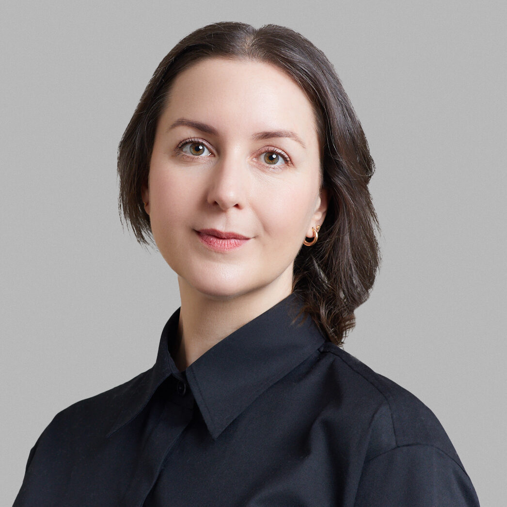 A woman with straight brown hair, wearing a black collared shirt, stands against a plain light gray background, looking calmly at the camera.