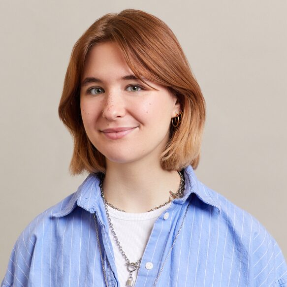 A young person with short, light brown hair smiles slightly. They wear a blue striped button-up shirt over a white top, layered necklaces, and small hoop earrings. The background is plain and light beige.