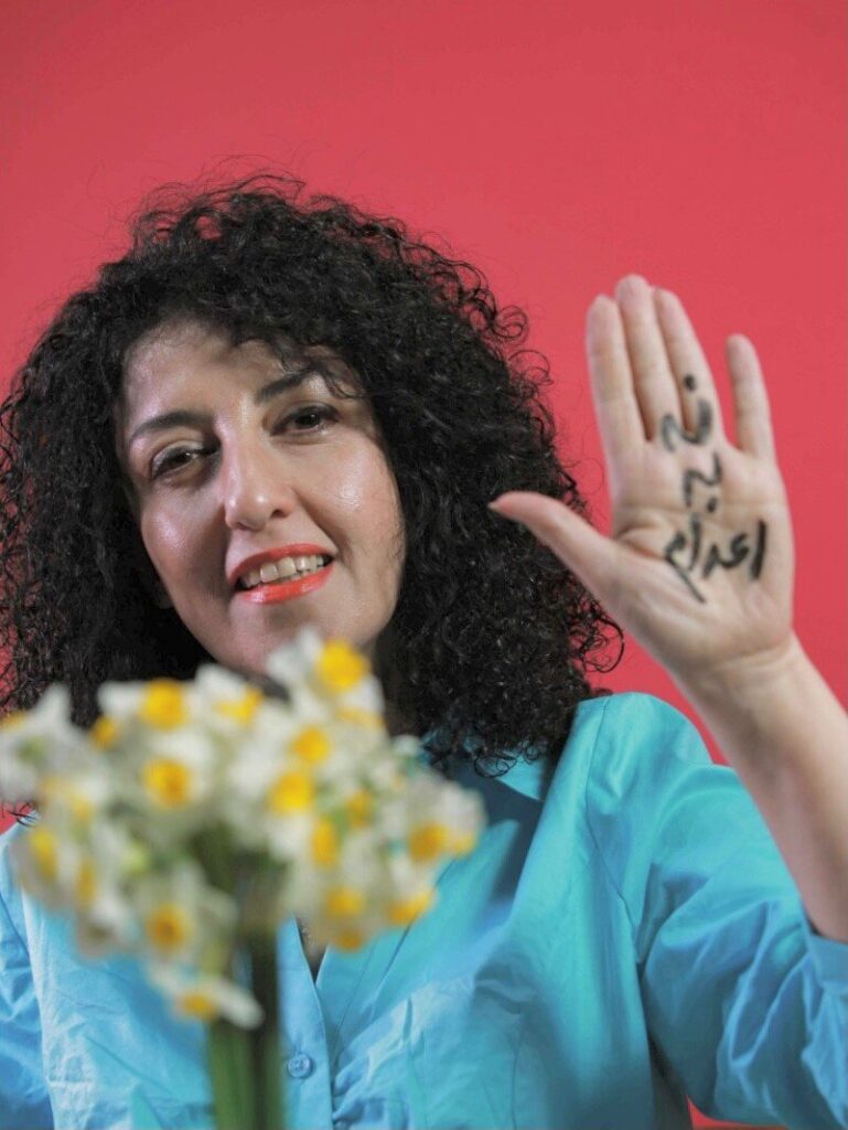 A woman with curly dark hair wearing a blue shirt smiles at the camera, holding up her hand with Arabic text written on it. She holds a bouquet of white and yellow flowers. The background is a solid pinkish-red color.