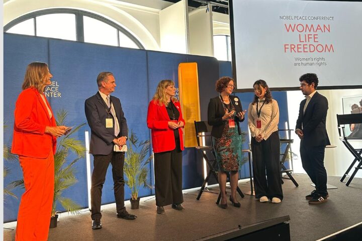 Six people stand on stage at the Nobel Peace Conference, with a screen behind them displaying "Woman Life Freedom" and "Women’s rights are human rights," as PEN America news highlights their voices for equality. Some speak while others listen attentively.