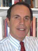 A middle-aged man with short dark hair, wearing a white and blue striped shirt and a red tie, is smiling in front of a bookshelf filled with books.