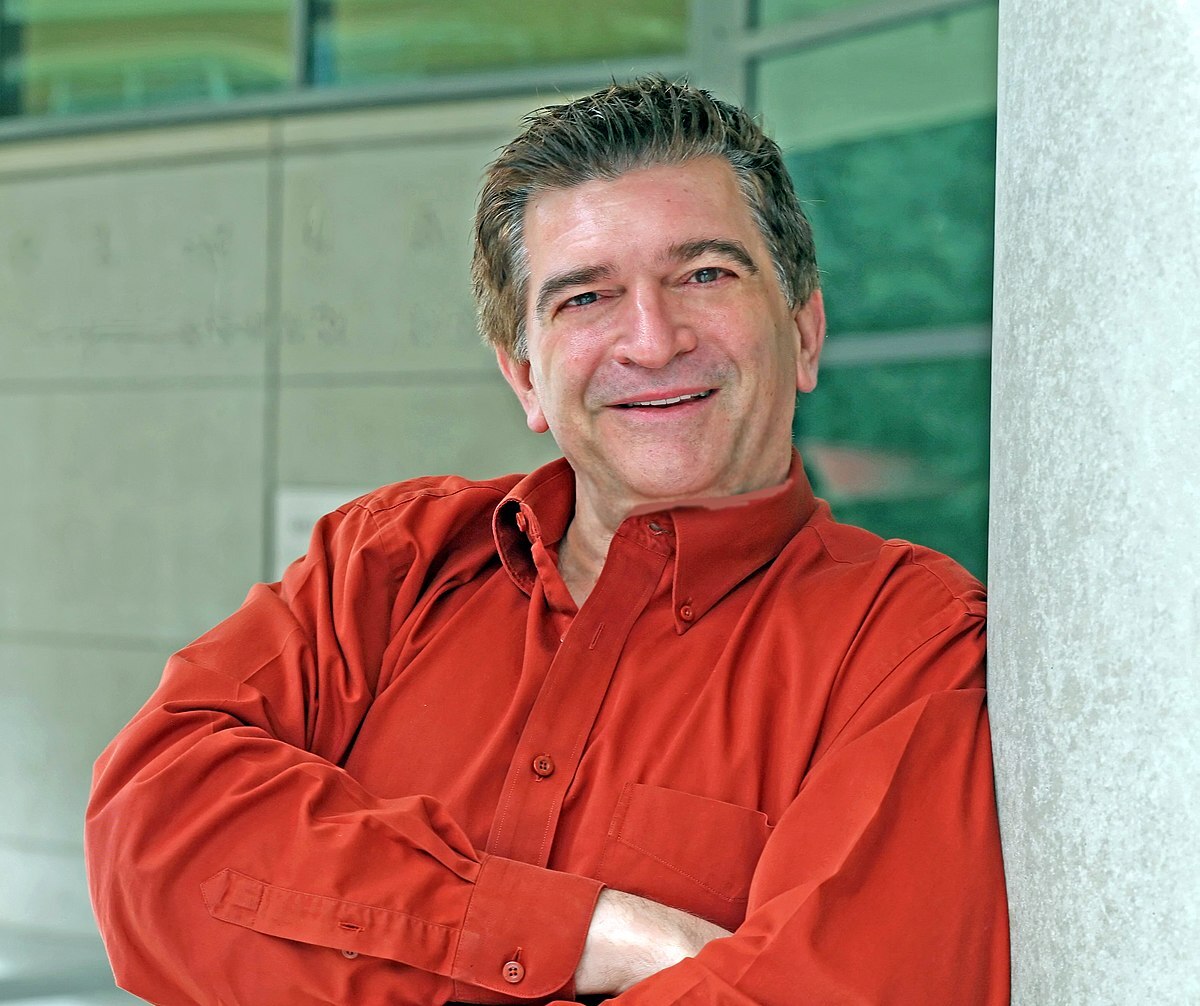 A middle-aged man with short brown hair, wearing a long-sleeved red shirt, stands smiling with arms crossed, leaning against a light-colored pillar in front of a modern building with glass windows.